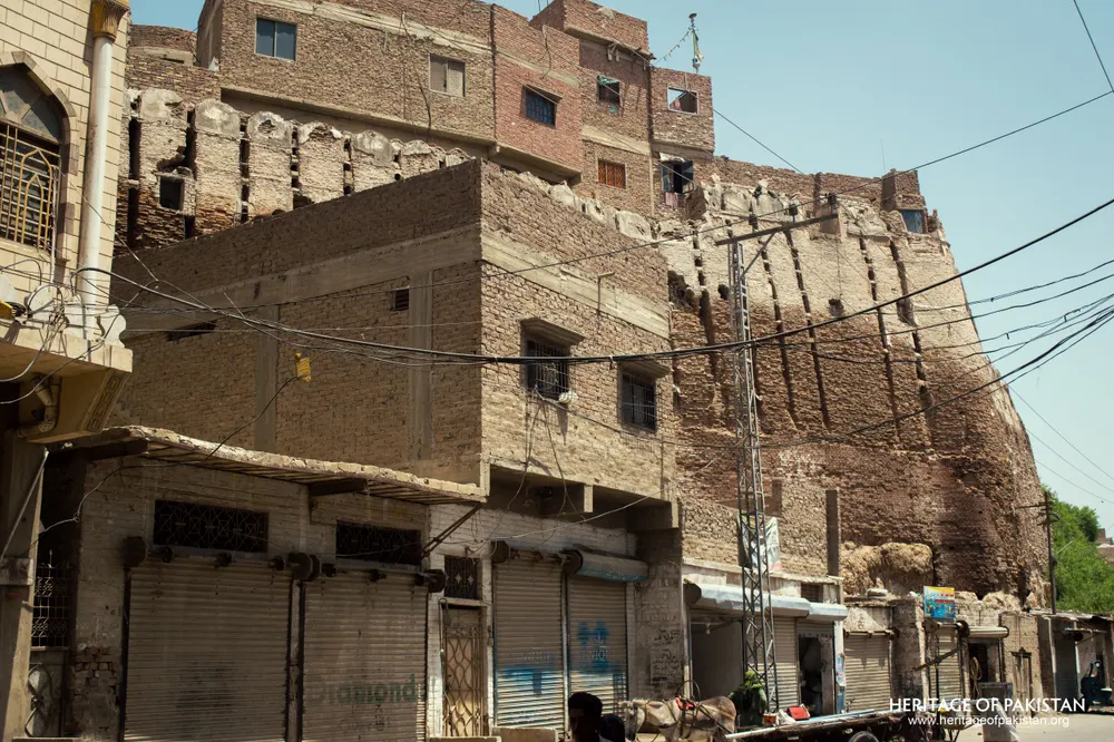 Street-level view of Pakka Qila's rampart rising above shuttered shopfronts and a parked cart