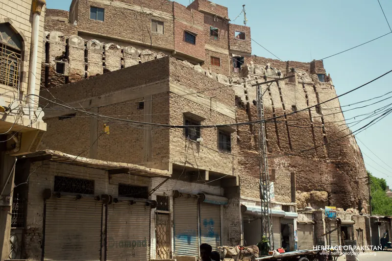 Street-level view of Pakka Qila's rampart rising above shuttered shopfronts and a parked cart