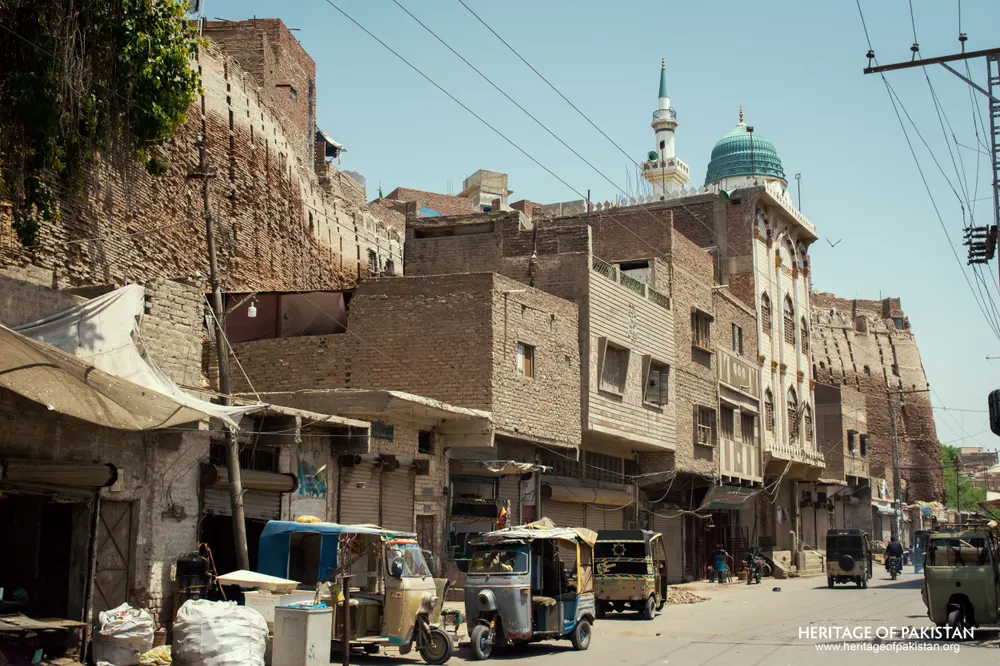Street-level view of Pakka Qila's rampart towering over a market row with a green-domed mosque