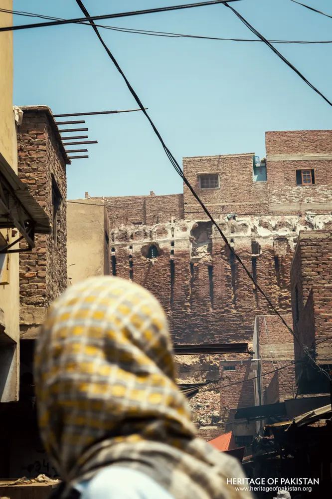 Distant view of Pakka Qila's ruined brick wall through a narrow alley, framed by a woman in a checkered headscarf